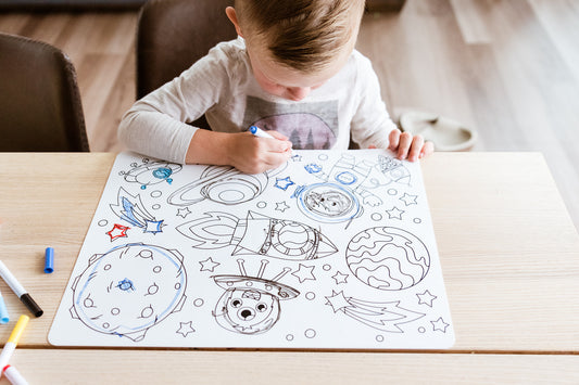 A young child sitting at a light wood table, drawing colorful space-themed illustrations like planets, rockets, and stars on a large white board with markers.