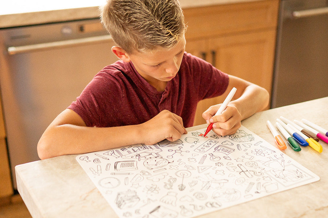 A young boy with short blond hair wearing a maroon t-shirt sits at a kitchen island, drawing on school themed reusable coloring mat with markers. Several colored markers are lined up on the counter next to him, with wooden kitchen cabinets and stainless steel appliances in the background.