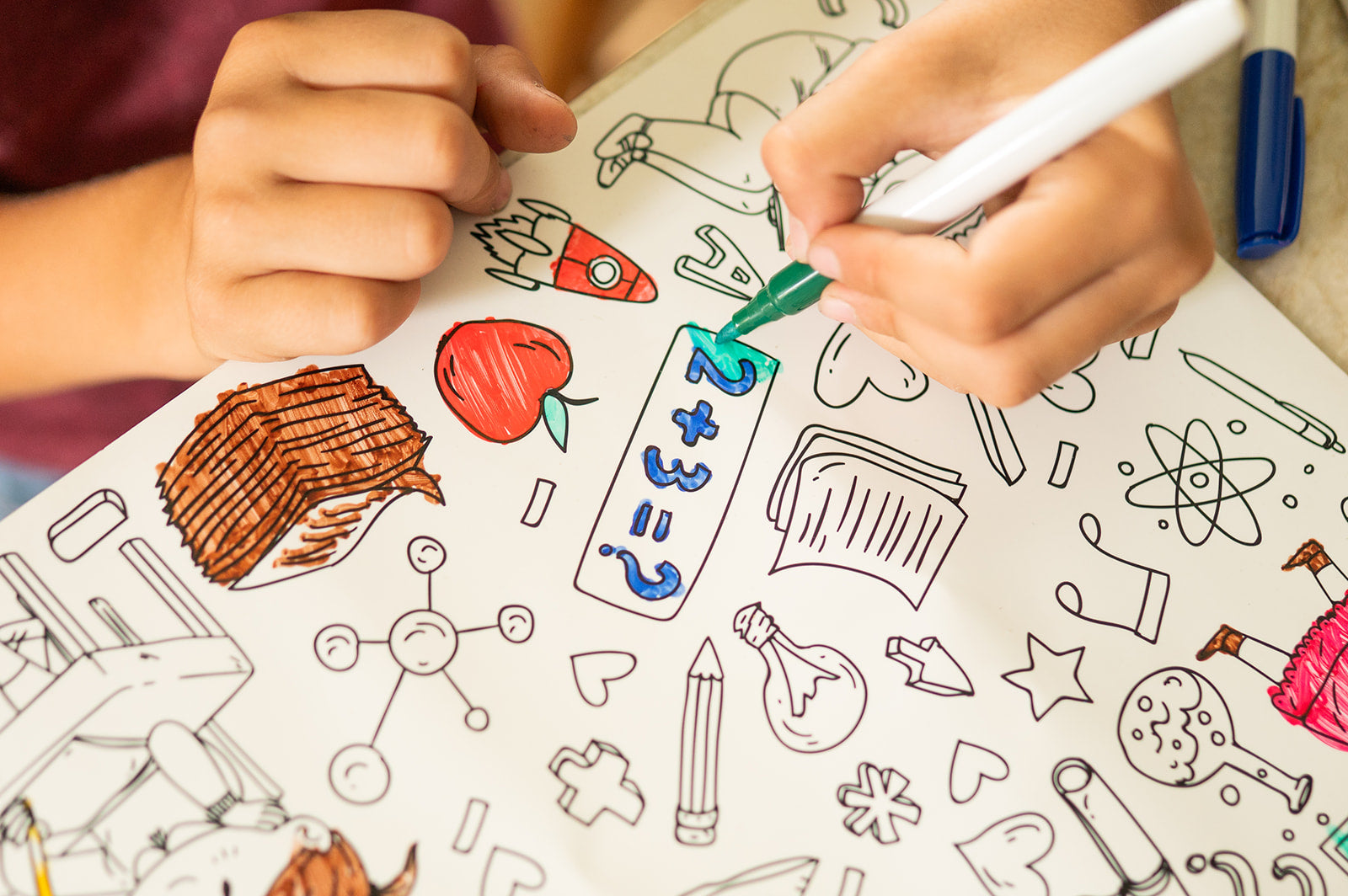 A close-up of a child's hands coloring School themed reusable silicone coloring mat with markers. The sheet features outlines of books, an apple, a rocket, and other school icons, with the child using a green marker.