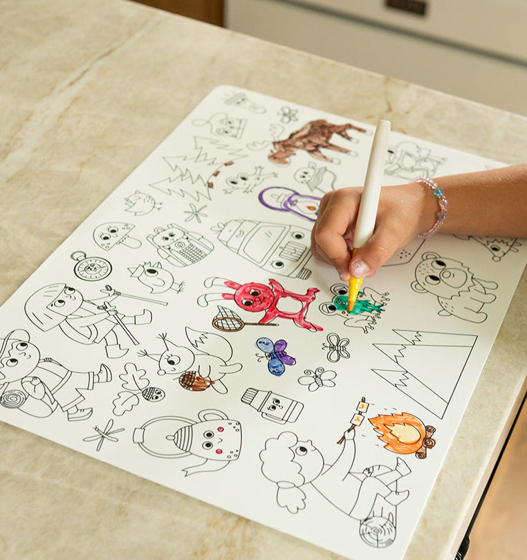 A child's hands are shown coloring a black and white cartoon drawing on a silicone placemat set on a kitchen counter. The placemat features various whimsical character illustrations.