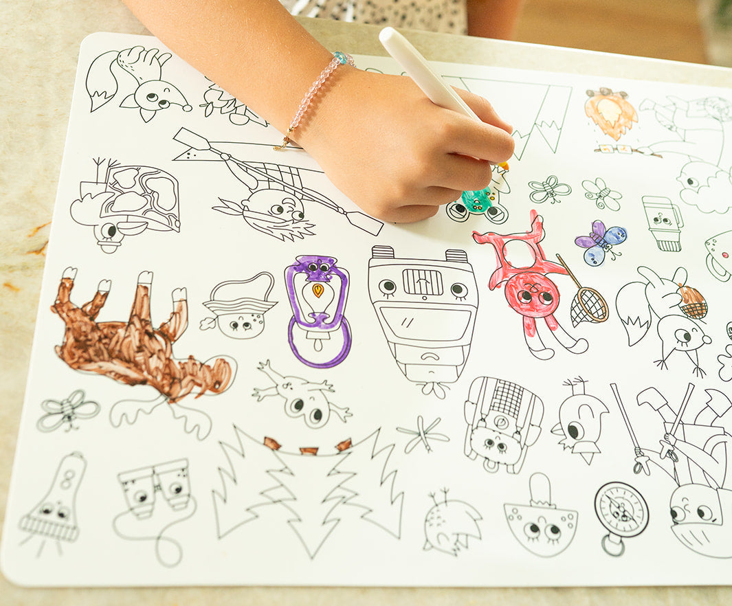 A young girl in a white polka-dot top draws on a large white magnetic board with a colorful marker, set on a light countertop with kitchen cabinets in the background.