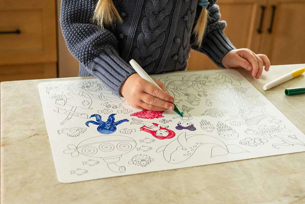 A young girl with braided hair sits at a kitchen counter, coloring a silicone coloring mat with markers.