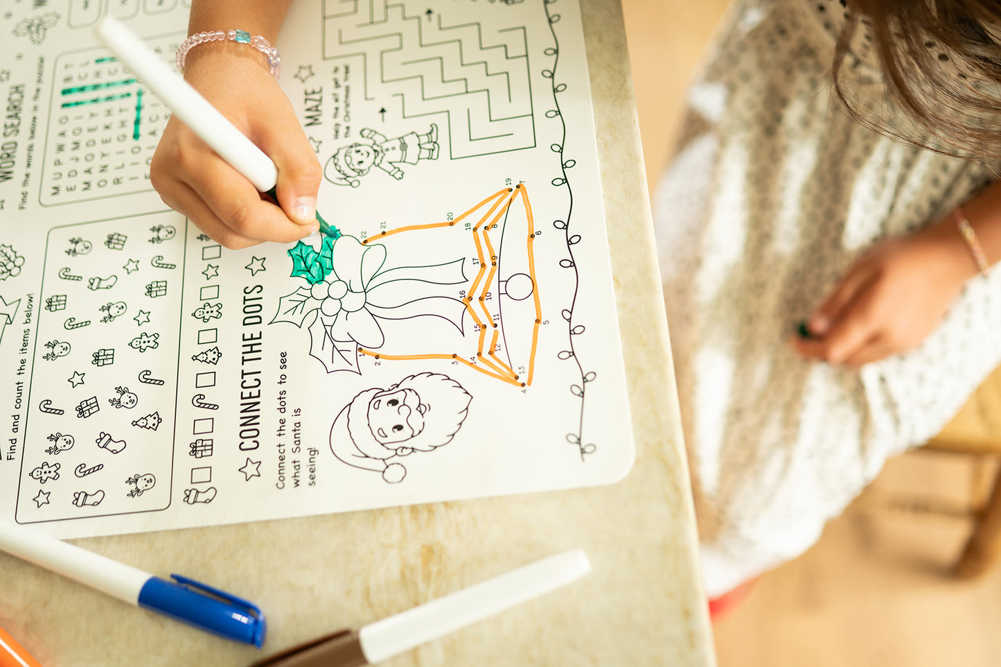 A child's hand holding a white marker, coloring a Christmas-themed worksheet on a light-colored desk. The worksheet features festive illustrations like Santa Claus and a gift box, along with other printable activities.