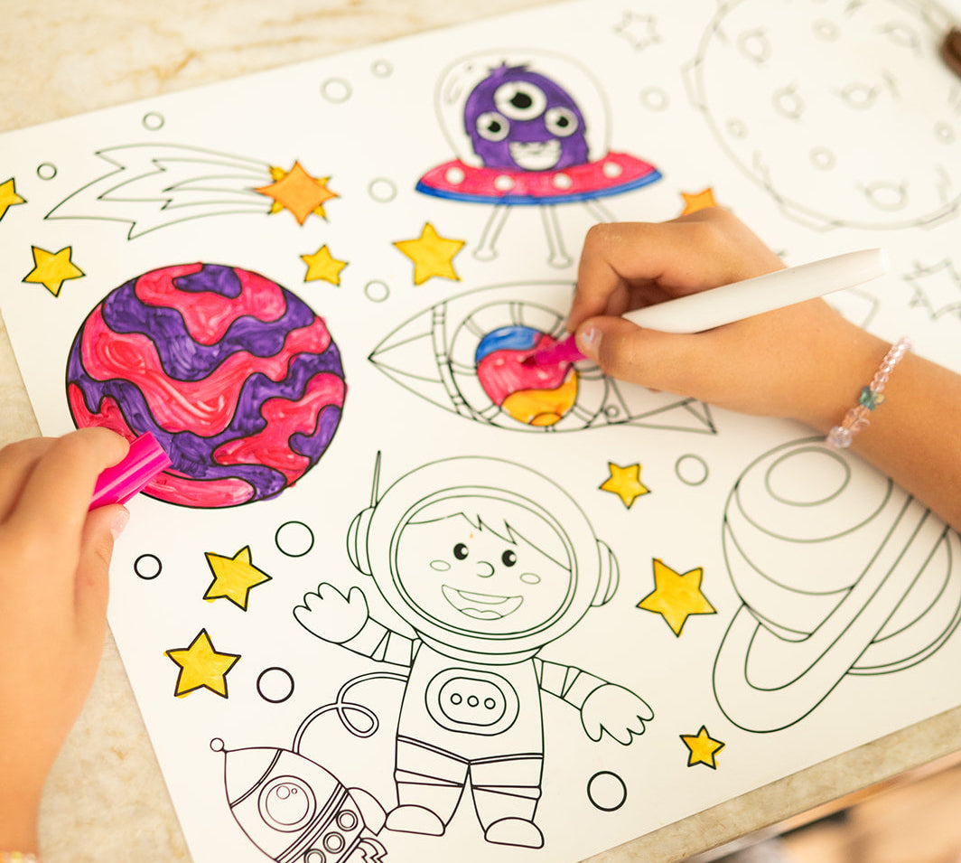 A child's coloring book featuring an outer space theme with a cartoon astronaut, planets, and stars, being colored with markers by two hands on a light-colored wooden table.
