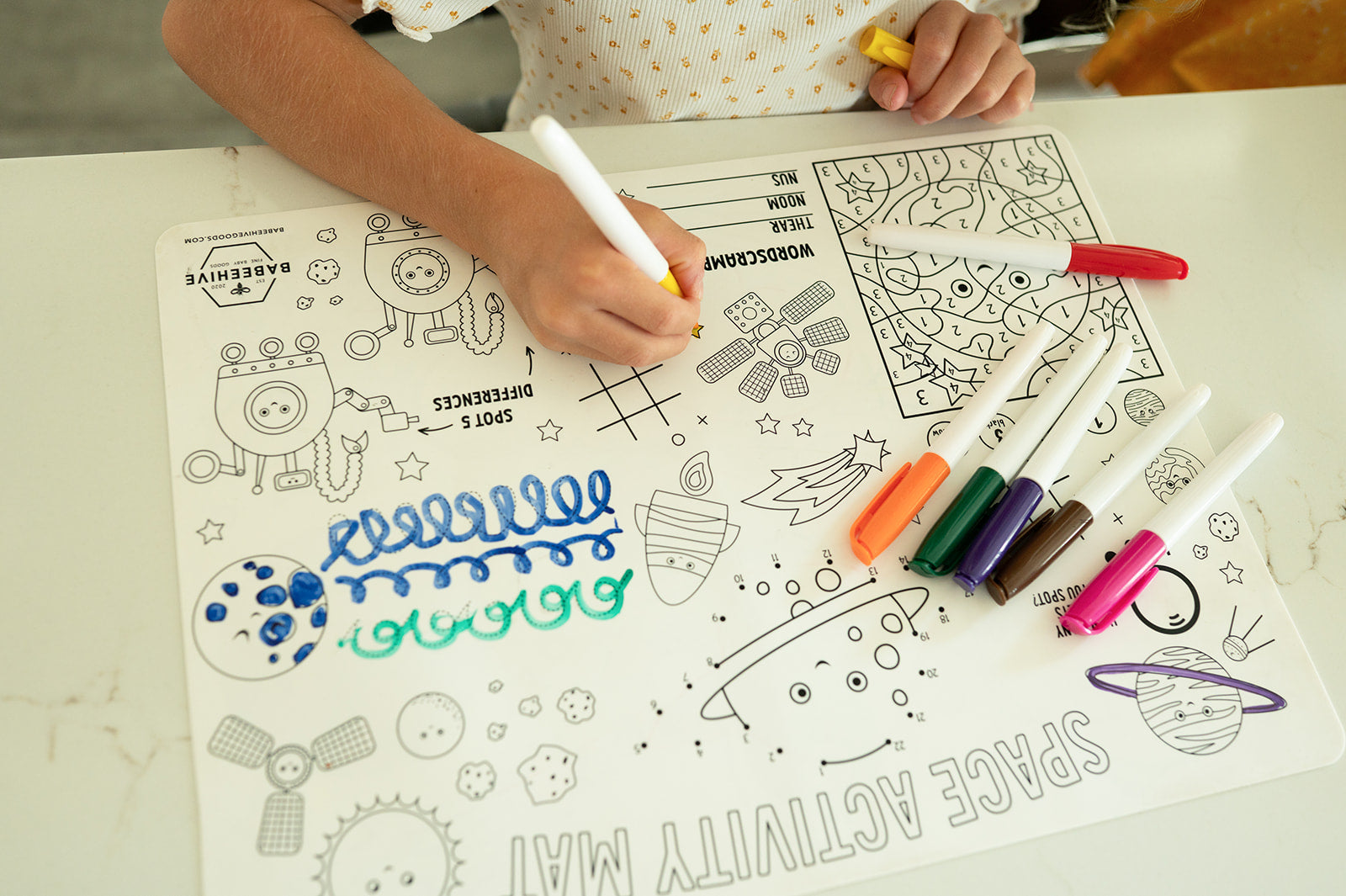 A child's hands drawing on a white space-themed activity mat with colorful markers, with the child's torso visible in the top left corner.