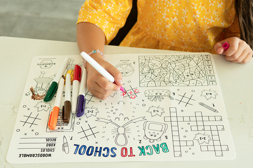A child in a yellow floral top sits at a white table, writing on a Back to School themed coloring mat with markers.