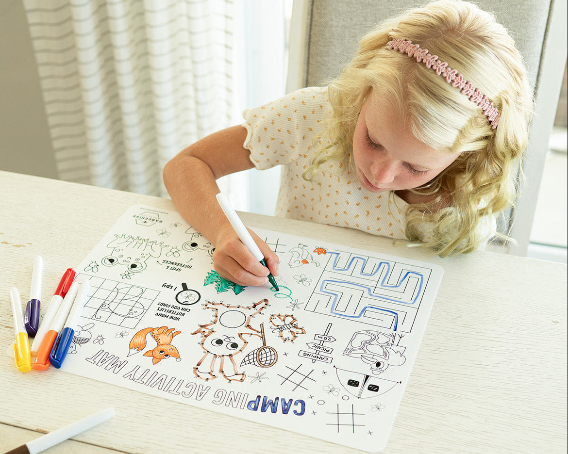 A young blonde girl with a pink headband sits at a light wood table, coloring in a camp-themed silicone coloring mat with markers. Several colored markers are scattered on the table next to her.