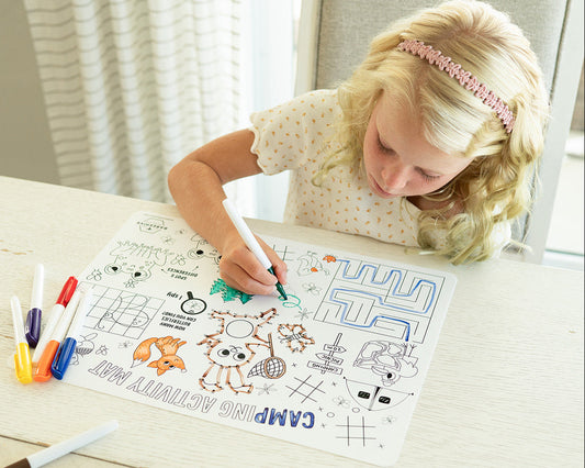A young blonde girl with a pink headband sits at a light wood table, coloring in a camp-themed silicone coloring mat with markers. Several colored markers are scattered on the table next to her.