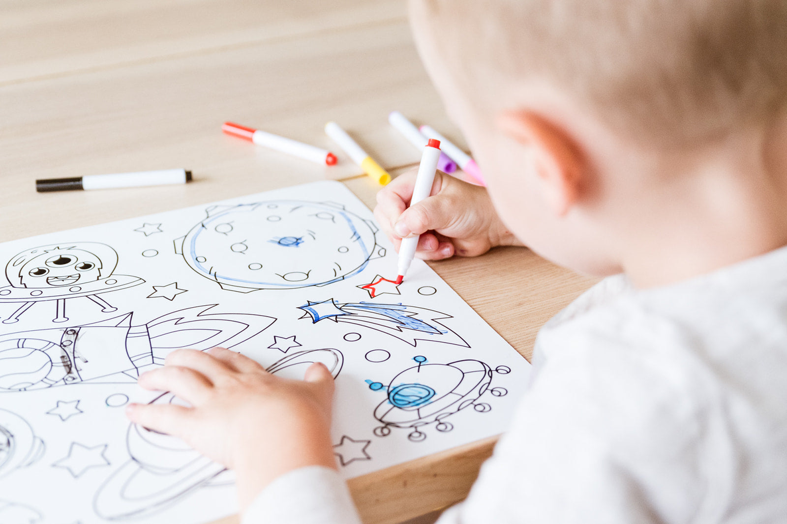 A child coloring a space silicone coloring mat with markers on a light wooden table.